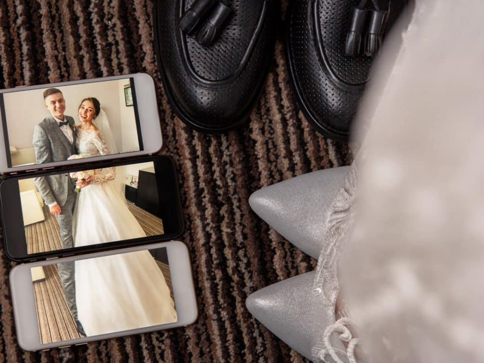 Fotografia slubna detali: buty pana i panny młodej obok smartfonów ze zdjęciami z ceremonii.