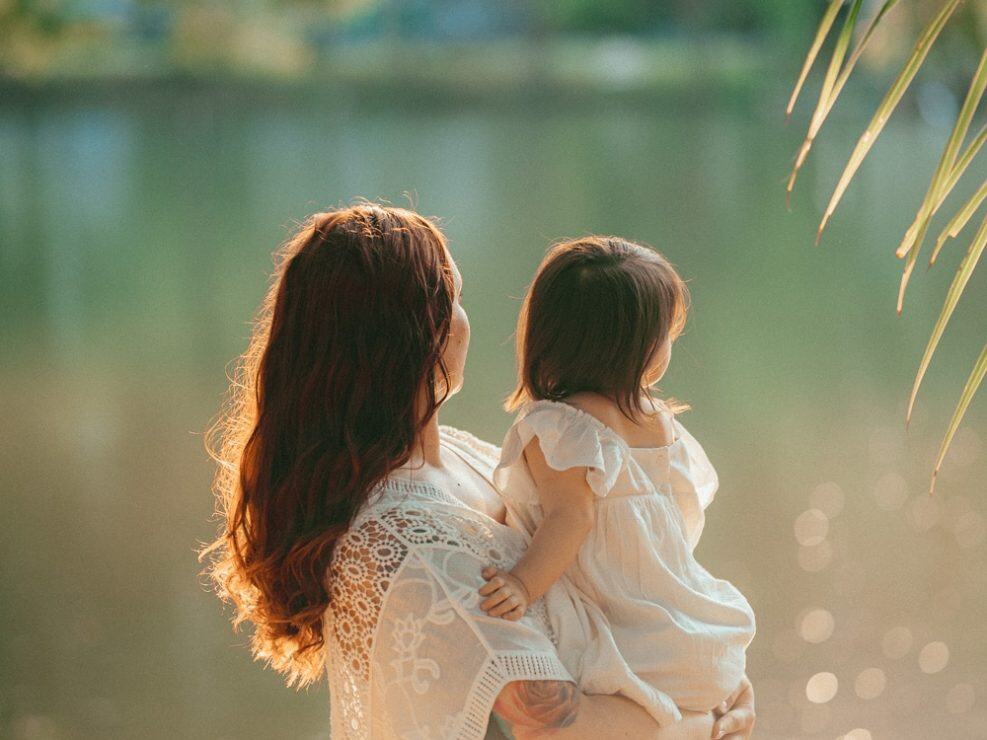 Reportaż chrzest: intymne zdjecia mamy z dzieckiem nad wodą w złotym świetle, ujęcie wykonane przez fotograf na chrzest.