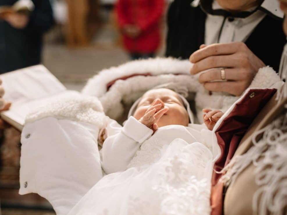 Reportaż chrzest warszawa: zbliżenie na niemowlę w szatce chrzcielnej podczas ceremonii w kościele. Kadr pełen wzruszenia, idealny jako zdjęcia chrzest na pamiątkę.