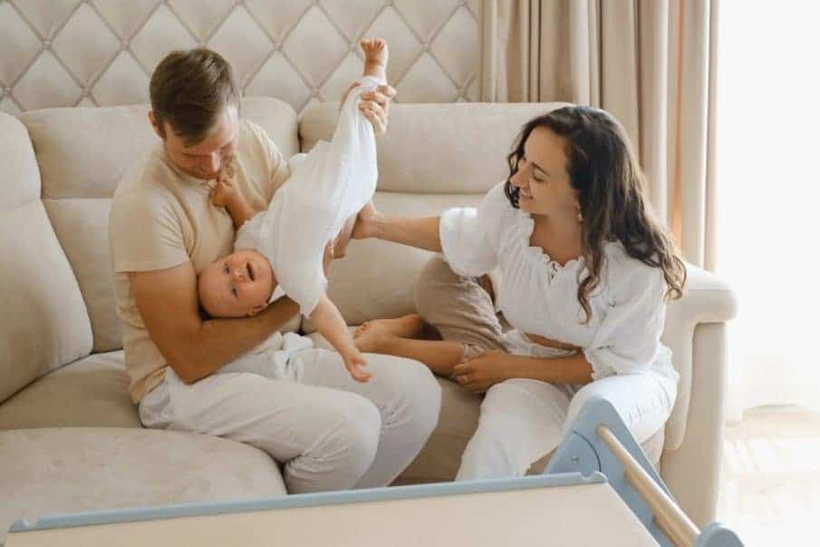 Fotograf chrzest warszawa: radosna rodzina bawi się z dzieckiem na sofie. Kadr z reportażu chrzcin u klienta, podkreślający szczęście i naturalność.