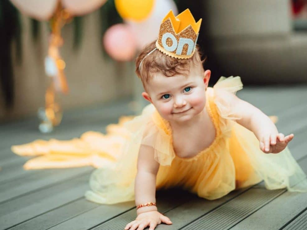 Fotograf roczek warszawa: uśmiechnięte dziecko w koronie urodzinowej i żółtej sukience. Sesja urodzinowa w stylu małej królewny, idealna pamiątka na roczek.