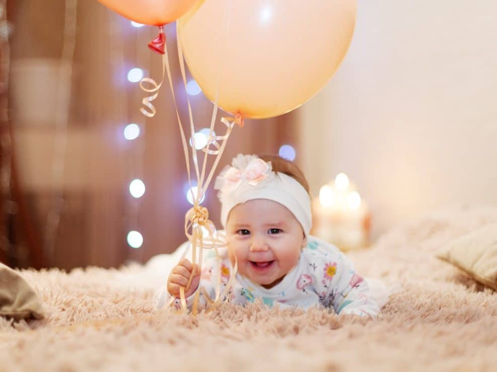 Sesja niemowleca warszawa: radosne, uśmiechnięte niemowlę na miękkim kocu, trzymające balony. Nastrojowe światło w tle.