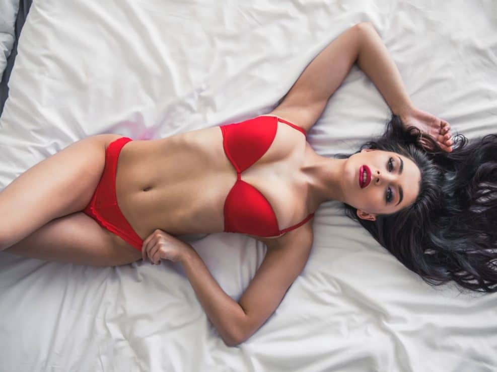 Sesja sensualna z góry: Brunetka w czerwonej bieliźnie na białej pościeli. Ujęcie top-down podkreślające kontrast kolorystyczny i zmysłowość.