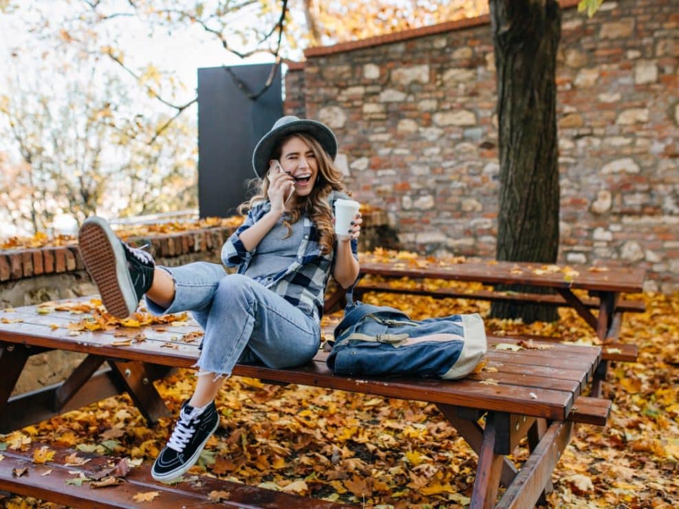 Sesja jesienna kobieca: uśmiechnięta kobieta rozmawia przez telefon, trzymając kawę w jesiennym plenerze. Zdjęcia jesienne lifestylowe.