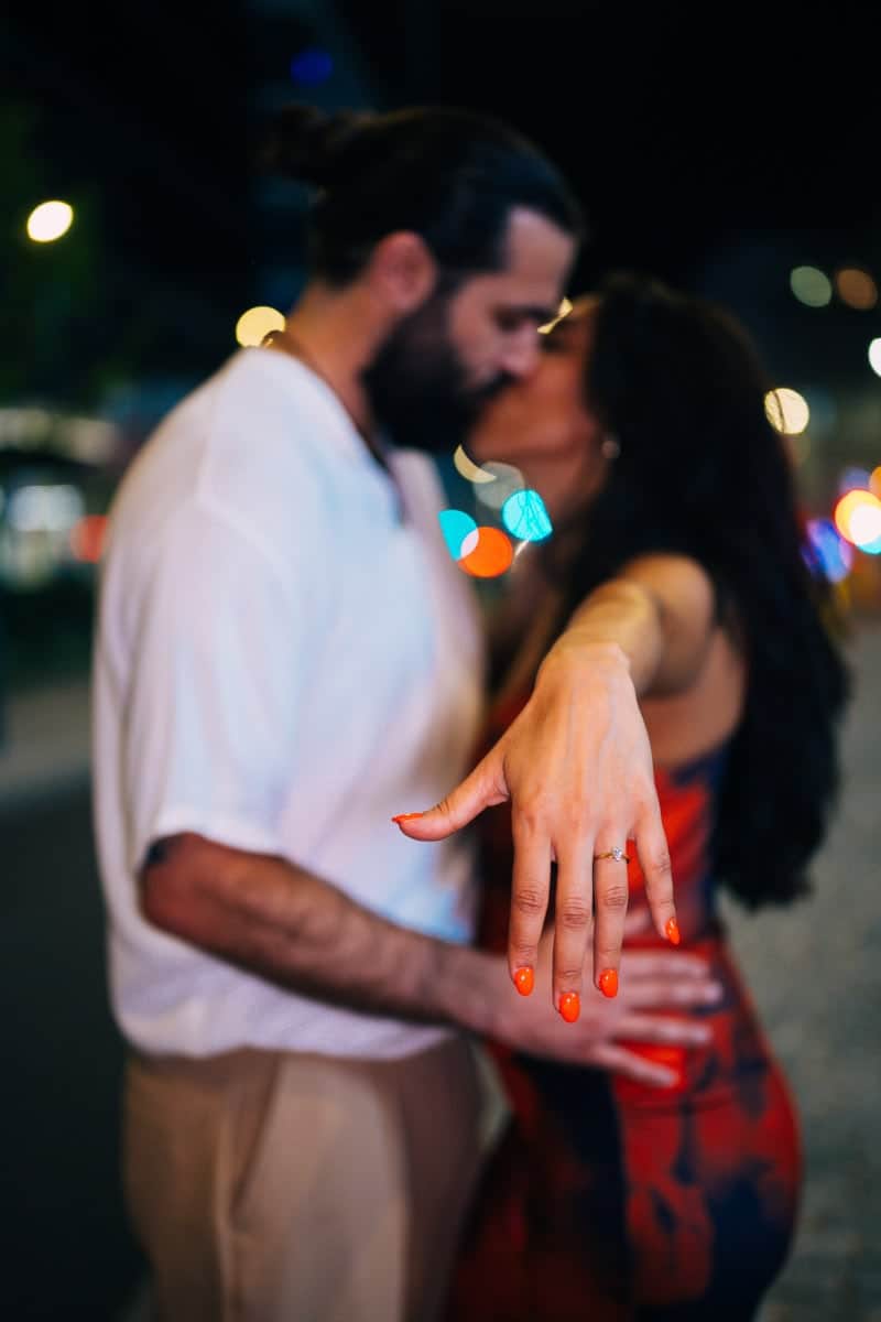 Romantyczna fotografia oświadczyn z pierwszym planem na pierścionek zaręczynowy na dłoni kobiety. Para w tle jest rozmazana (bokeh), całując się. Zdjęcie z nocnej sesji zaręczynowej w warszawie, z kolorowymi światłami miejskimi.