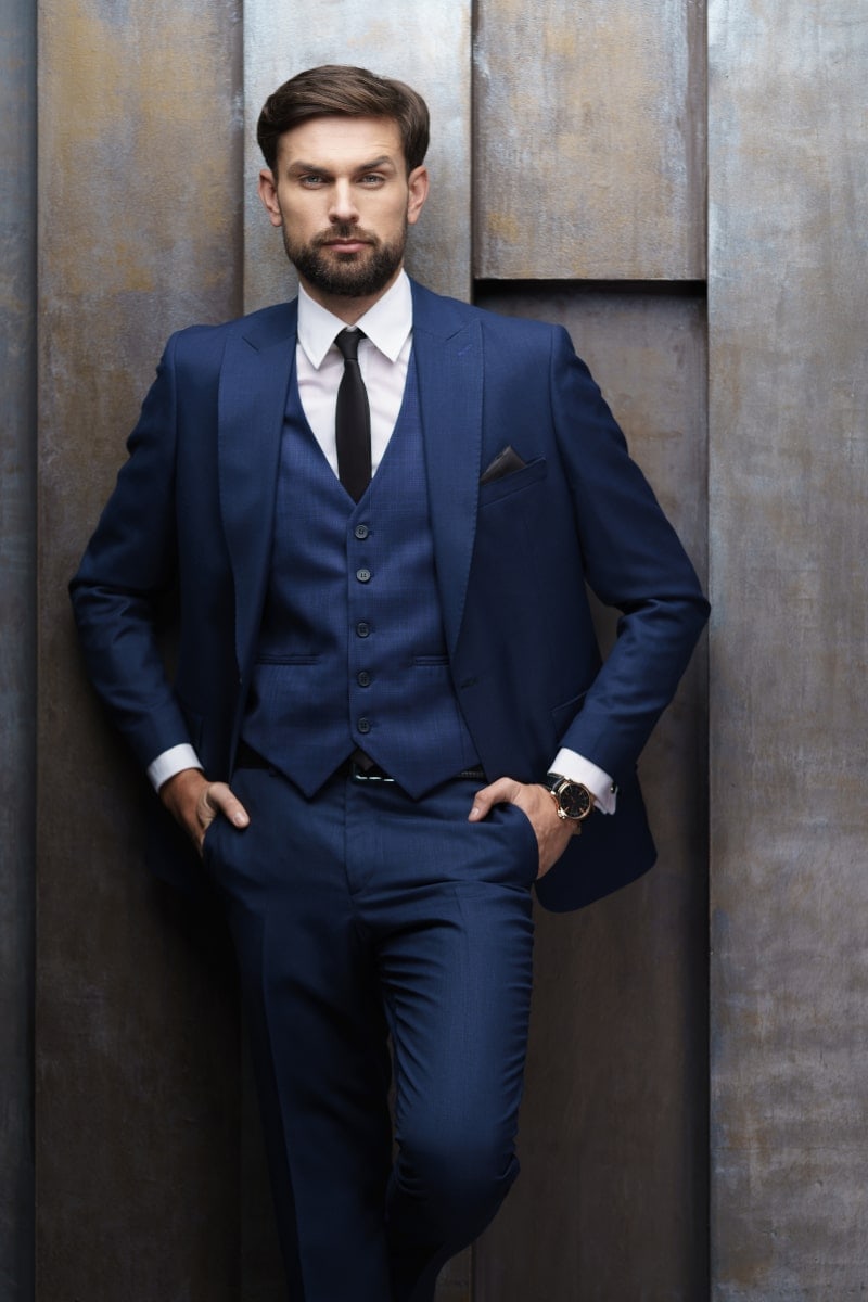 Elegancki, profesjonalny portret biznesowy mężczyzny w granatowym garniturze, kamizelce i czarnym krawacie. Zdjęcie wykonane w studio na tle nowoczesnej, strukturalnej ściany. Idealny portret dla kadry menadżerskiej i prezesów.