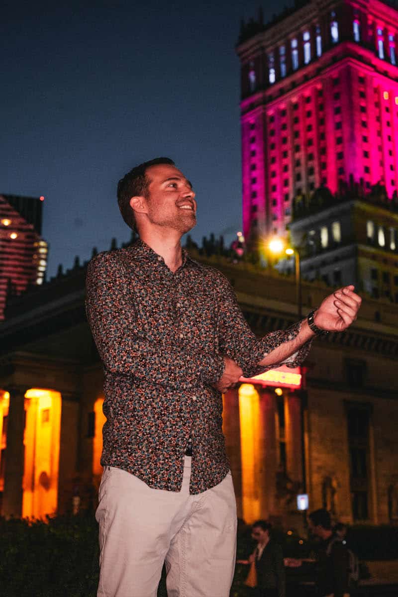 Nocny portret mężczyzny z imprezy w centrum warszawy. Uśmiechnięty mężczyzna w kwiecistej koszuli, z podwiniętymi rękawami, pozuje na tle rozświetlonego neonami pałacu kultury i nauki. Zdjęcie lifestylowe z nocnego życia warszawy.