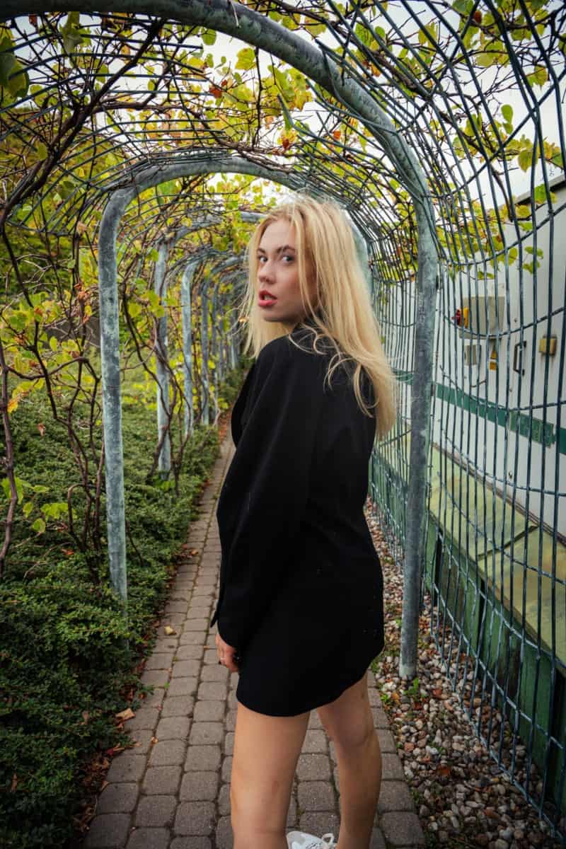 Portret z sesji kobiecej wykonany w ogrodzie biblioteki uniwersytetu warszawskiego. Blondynka w czarnej marynarce pozuje w tunelu z zielonymi pnączami i ogrodzeniem. Sesja lifestylowa w pięknej, naturalnej scenerii warszawy.