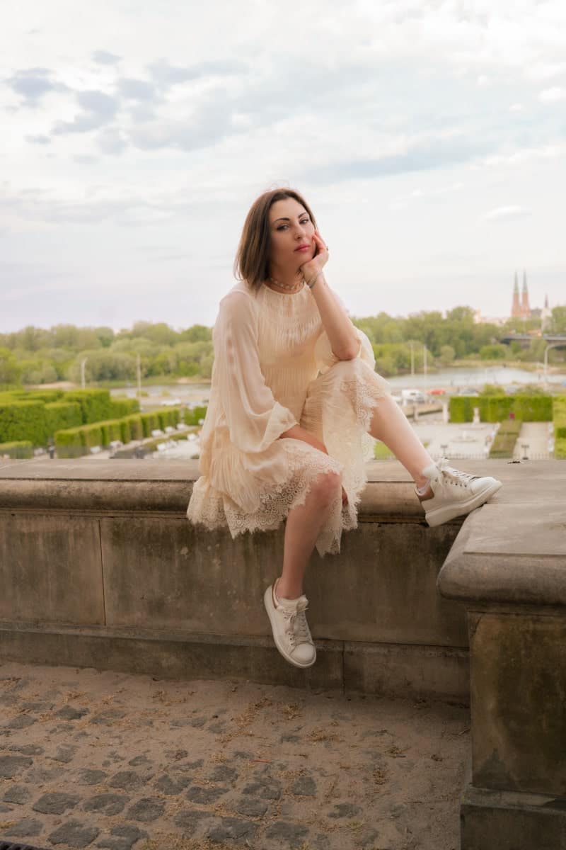 Portret z sesji kobiecej w letniej sukience i białych sneakersach, siedzącej na tle ogrodów zamku królewskiego w warszawie. Elegancka kobieta pozuje na tarasie widokowym z panoramą wisły i pragi w tle. Zdjęcie lifestylowe wykonane przez fotografa z warszawy.