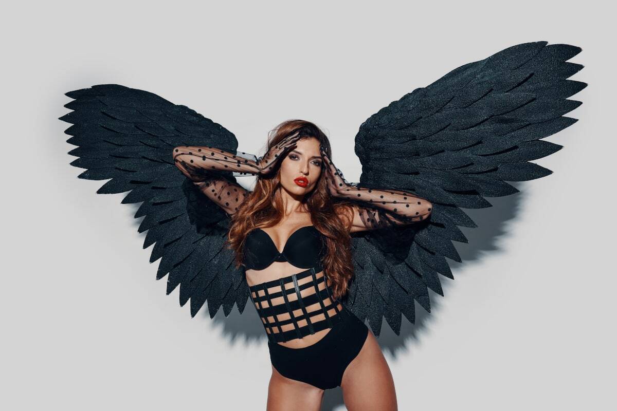 Fotograf sensualny. Artystyczna sesja zdjęciowa w studio. Kobieta w czarnej bieliźnie i dużych, czarnych skrzydłach. Neutralne tło i profesjonalne oświetlenie. Zdjęcie ilustruje kreatywne i kontrolowane możliwości sesji w studio.