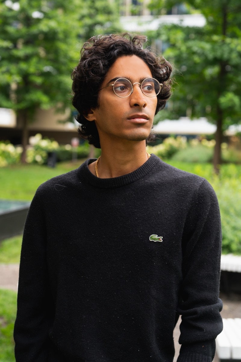 Portret młodego mężczyzny w okularach w cienkiej oprawie i czarnym swetrze, wykonany w parku w warszawie. Zdjęcie z sesji tinder/randkowej, idealne do profilu, z naturalnym tłem zielonej roślinności i miejskiej architektury.