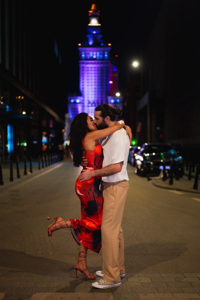 Nocna sesja zaręczynowa na ulicy w centrum warszawy. Para całuje się namiętnie w wieczornym świetle, kobieta w długiej, czerwonej sukience i szpilkach. Romantyczne zdjęcie zaręczynowe w mieście, wykonane przez fotografa z warszawy.