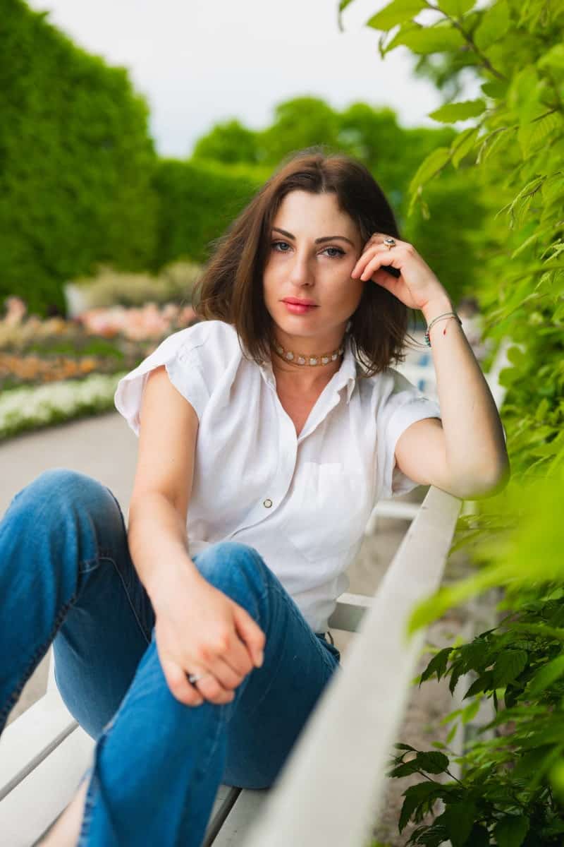 Portret kobiety w białej, lnianej koszuli i jeansach, siedzącej na ławce w ogrodach królewskich w warszawie. Sesja zdjęciowa w plenerze z intensywnie zielonym, wiosennym tłem. Ujęcie lifestylowe wykonane przez fotografa z warszawy.