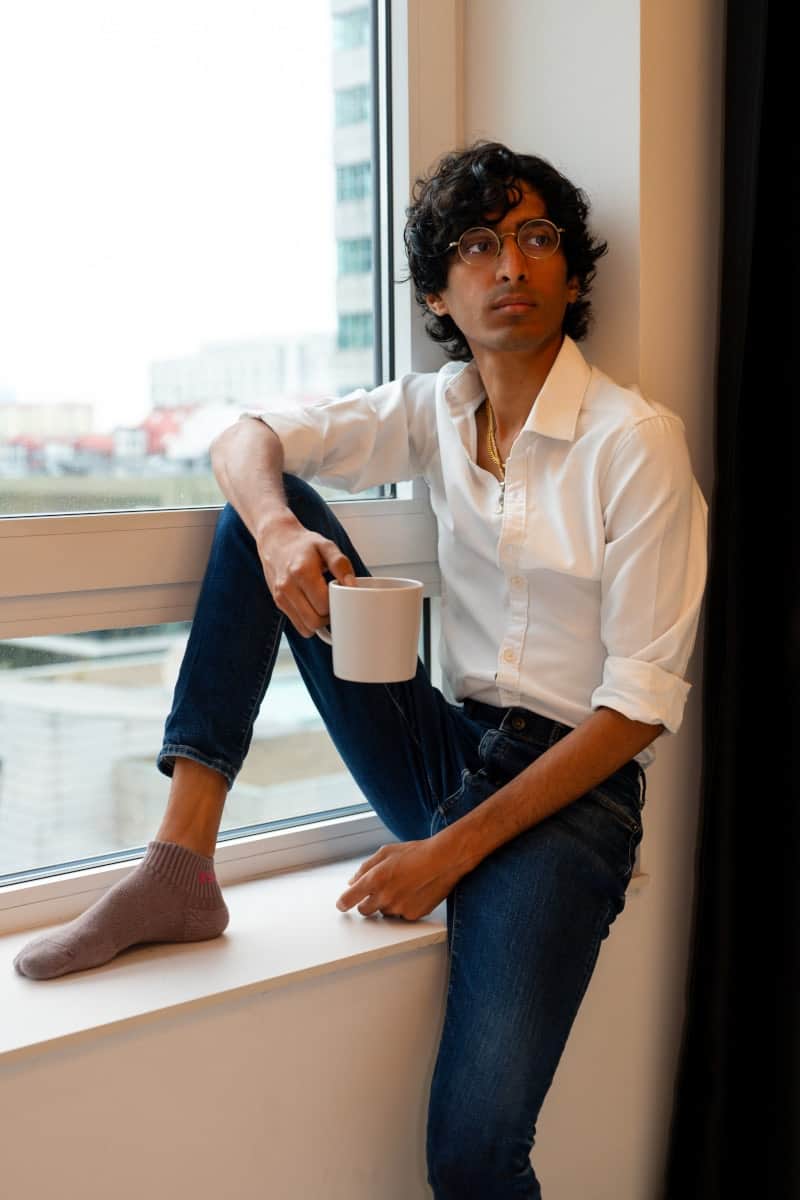 Lifestylowe zdjęcie do profilu tinder. Mężczyzna w okularach w cienkiej oprawie, białej koszuli i jeansach siedzi na parapecie nowoczesnego apartamentu, trzymając kubek kawy. Portret w stylu kawalerki z widokiem na miasto, wykonany przez fotografa z warszawy.