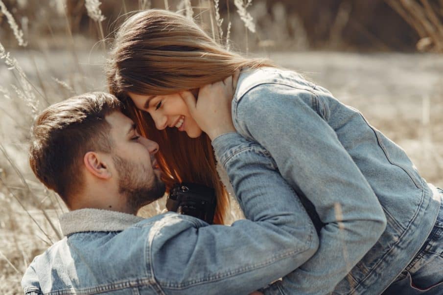 Uśmiechnięta para w denimowych kurtkach przytula się czule na tle słonecznego, wysuszonego pola lub łąki. Kobieta ma długie włosy i dotyka twarzy mężczyzny, który trzyma aparat. Zdjęcie ilustruje ofertę vouchera na sesję fotograficzną dla par i sugeruje zapytanie o cenę.