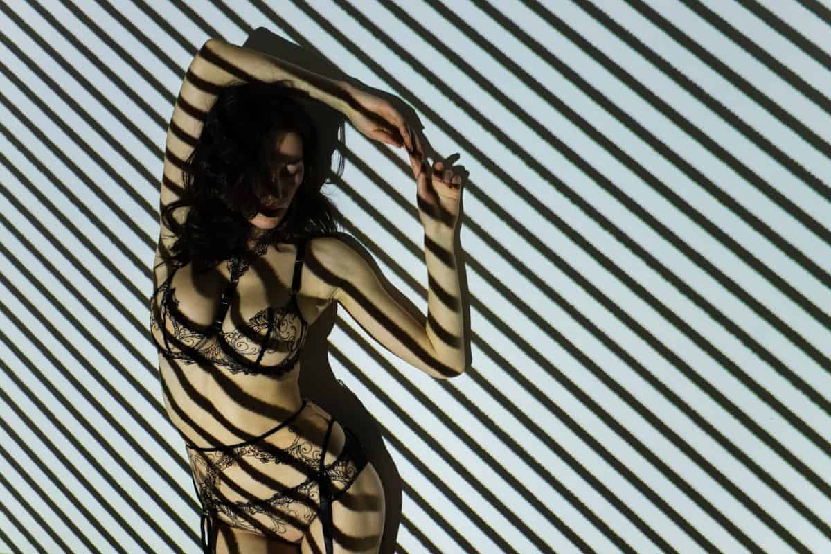 Fotograf sensualny. Artystyczna sesja zdjęciowa. Kobieta w bieliźnie (gorset) na ciemnym tle, oświetlona kontrolowanym światłem dającym wzór w paski. Zdjęcie ilustruje wyrafinowane "efekty świetlne w studio" do sesji sensualnej.