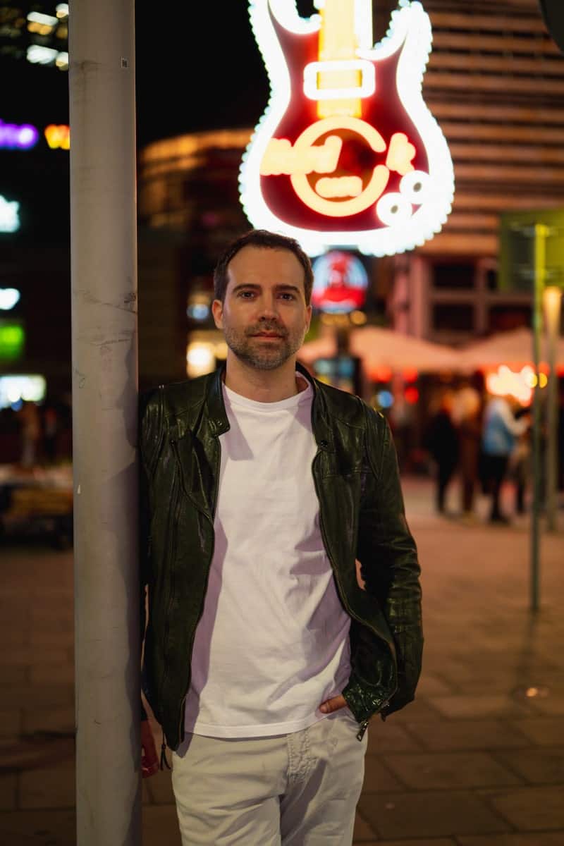 Nocne zdjęcie do tindera przedstawiające mężczyznę w czarnej skórzanej kurtce i białej koszulce, opartego o słup. Tło to rozmyte, neonowe światła lokali w centrum warszawy, w tym logo hard rock cafe. Męski portret lifestyle'owy z nocnej warszawy.