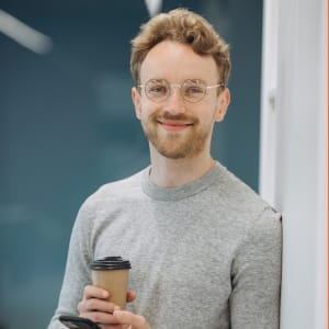 Uśmiechnięty mężczyzna w okularach i szarym swetrze, trzymający kawę na wynos; naturalny portret lifestylowy wykonany w nowoczesnym wnętrzu.