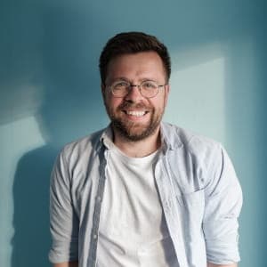 Zadowolony klient polecający usługi fotograficzne; uśmiechnięty mężczyzna w okularach i błękitnej koszuli na jasnoniebieskim tle.