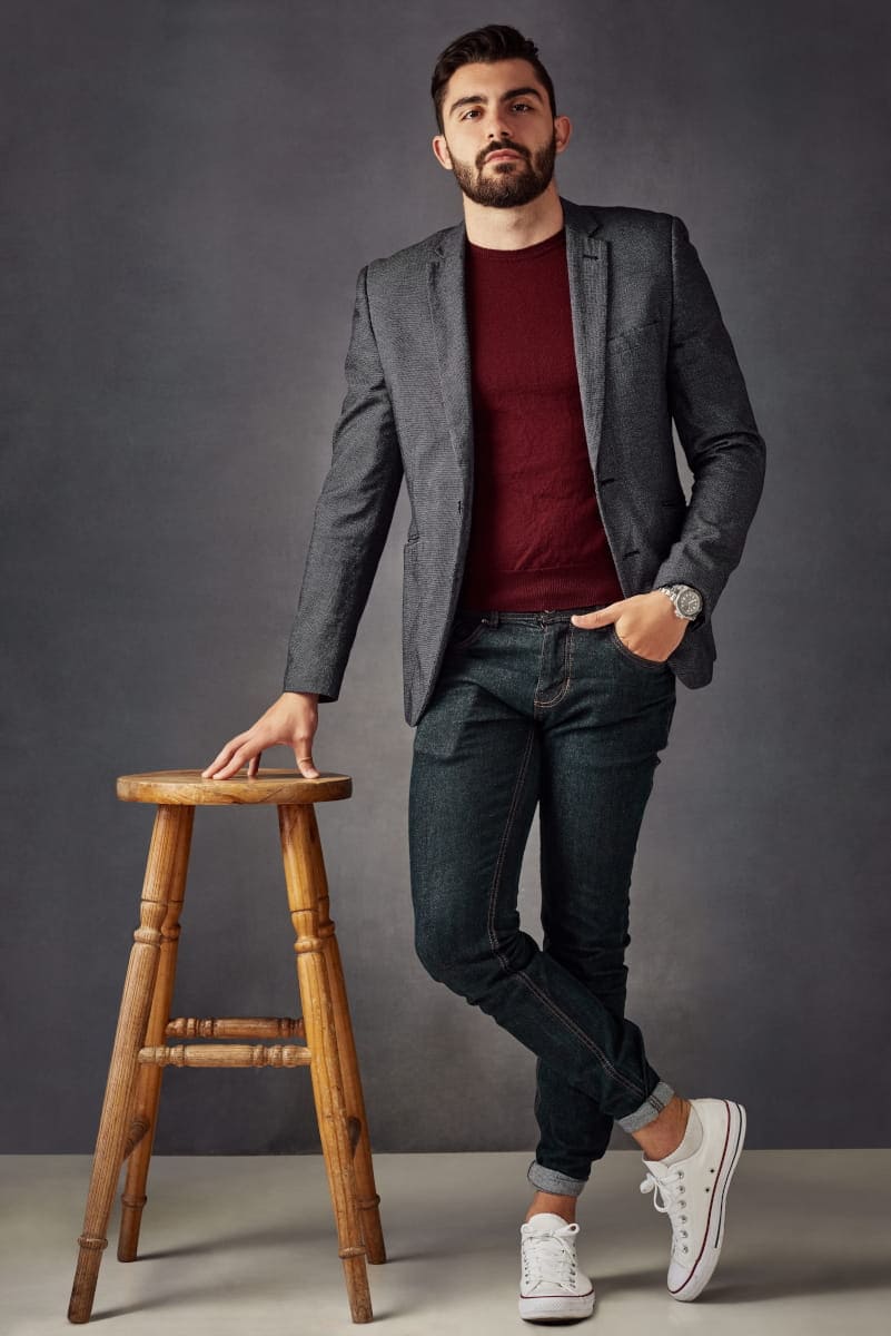 Mężczyzna w szarej marynarce, bordowym swetrze i jeansach pozujący przy drewnianym hokerze – profesjonalna sesja zdjęciowa na tinder w warszawie.