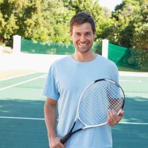 Uśmiechnięty mężczyzna w jasnoniebieskim t-shircie trzymający rakietę tenisową na korcie; naturalny kadr pokazujący aktywne spędzanie czasu.