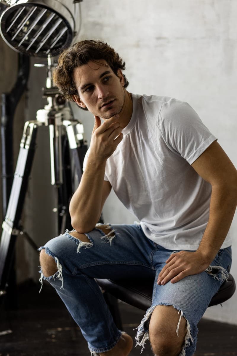 Mężczyzna w białym t-shircie i przecieranych dżinsach siedzący w studio fotograficznym, pozujący w zamyślony sposób na tle profesjonalnej lampy.