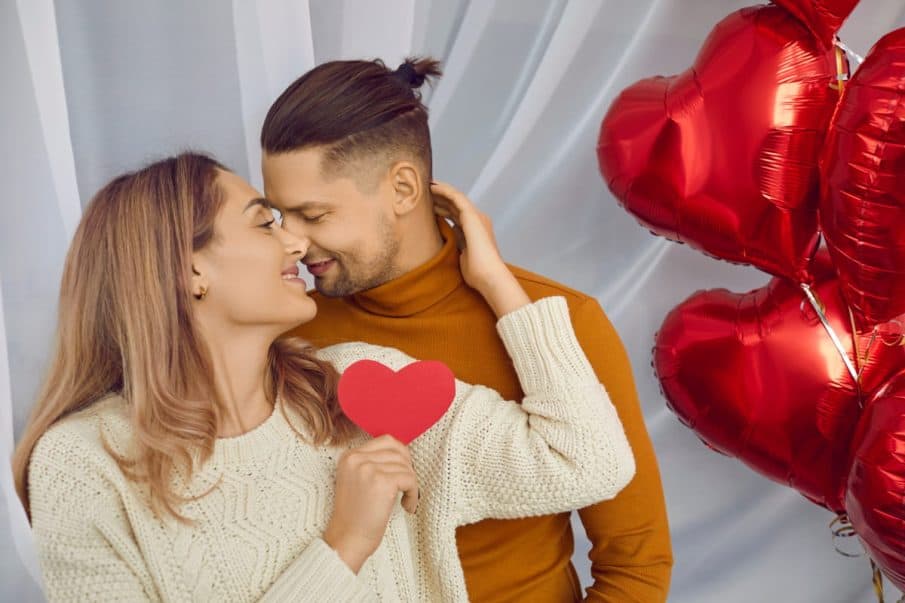 Para w przytulnych swetrach – kobieta trzymająca papierowe czerwone serce i mężczyzna czule stykający się z nią nosami podczas sesji zdjęciowej dla par w warszawie na tle czerwonych balonów