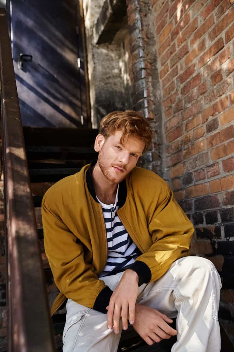 Mężczyzna w żółtej kurtce bomberce i pasiastej koszulce siedzący na zewnętrznych schodach w warszawie – naturalne zdjęcie na profil randkowy.
