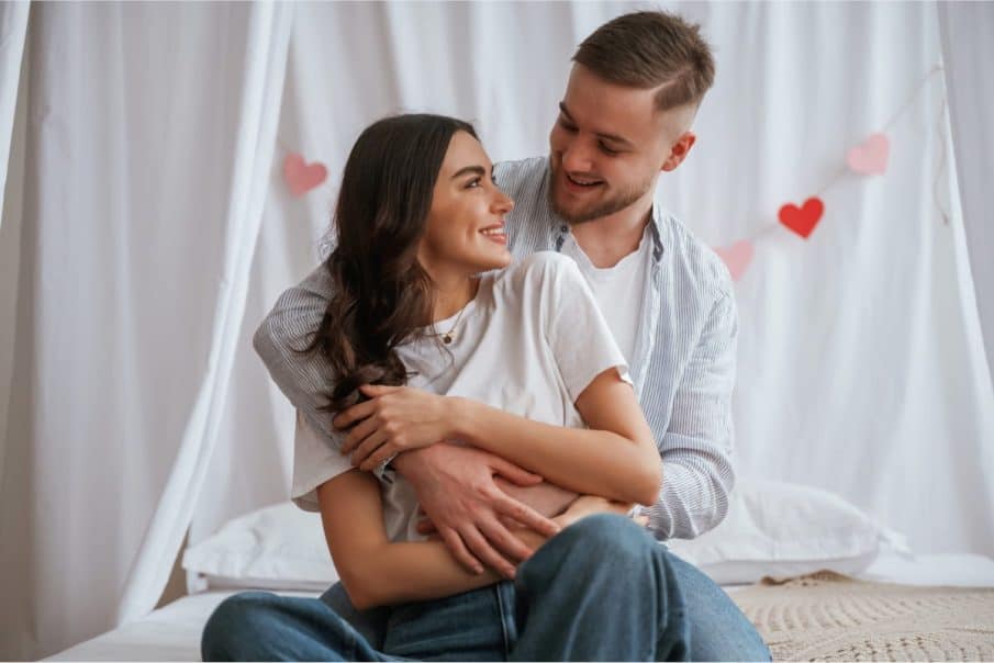 Zakochana para przytulająca się na łóżku podczas sesji na walentynki w warszawie; kobieta i mężczyzna patrzą na siebie z uśmiechem w jasnym, romantycznym wnętrzu udekorowanym girlandą z serc.