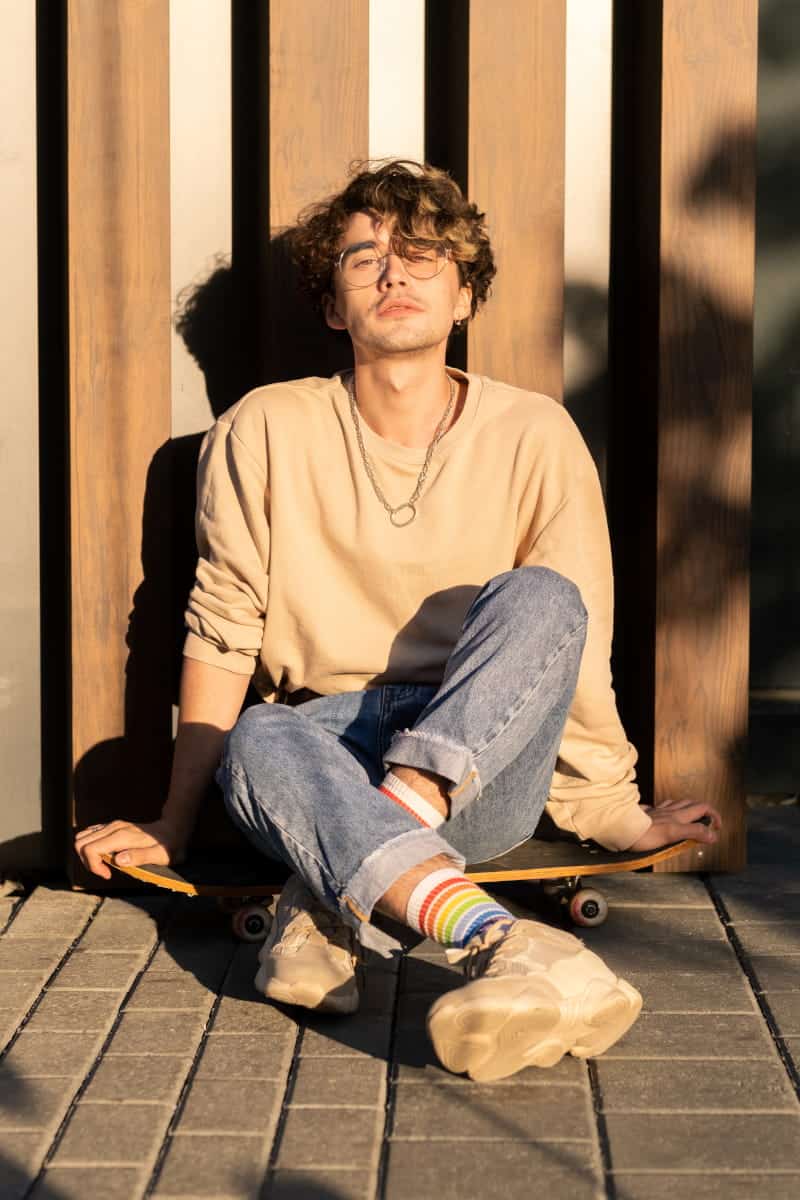 Młody mężczyzna w okularach i beżowej bluzie, siedzący na deskorolce w słońcu. Naturalna sesja zdjęciowa typu lifestyle na profil randkowy i instagram.