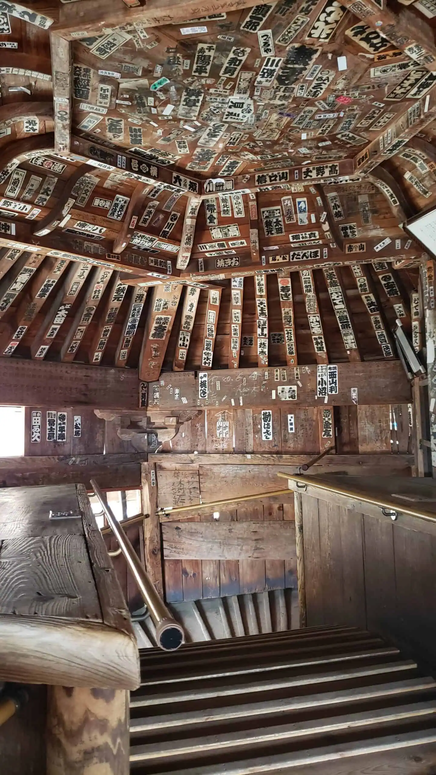 A wooden interior featuring a steep staircase leading to a room with a high, textured ceiling. The ceiling and walls are adorned with numerous senjafuda labels, creating a rustic and patterned aesthetic. Rails are installed along the staircase for support.