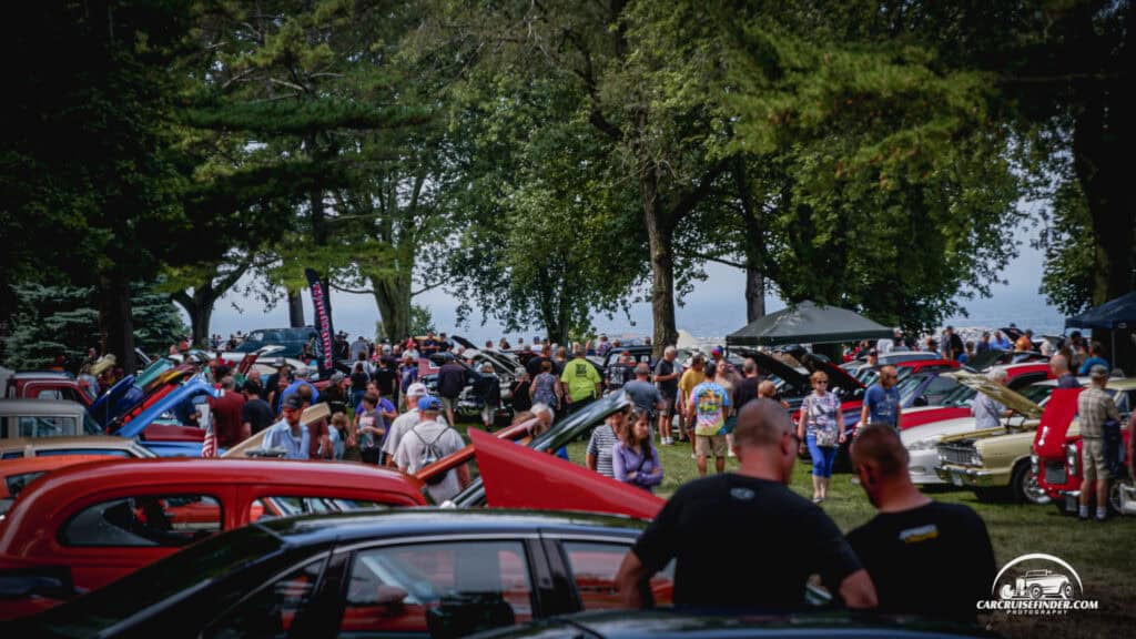 Crowd of visitors walking among classic cars under shade trees at a car show