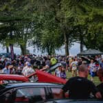 Crowd of visitors walking among classic cars under shade trees at a car show
