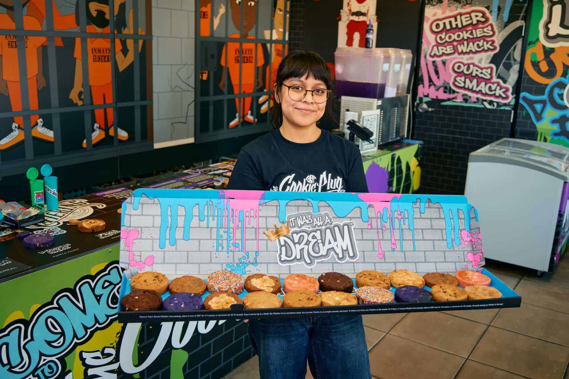 Girl with bangs and glasses holding a very large catering box of cookies that are thick and in many different kinds of flavors in front of some colorful graffiti