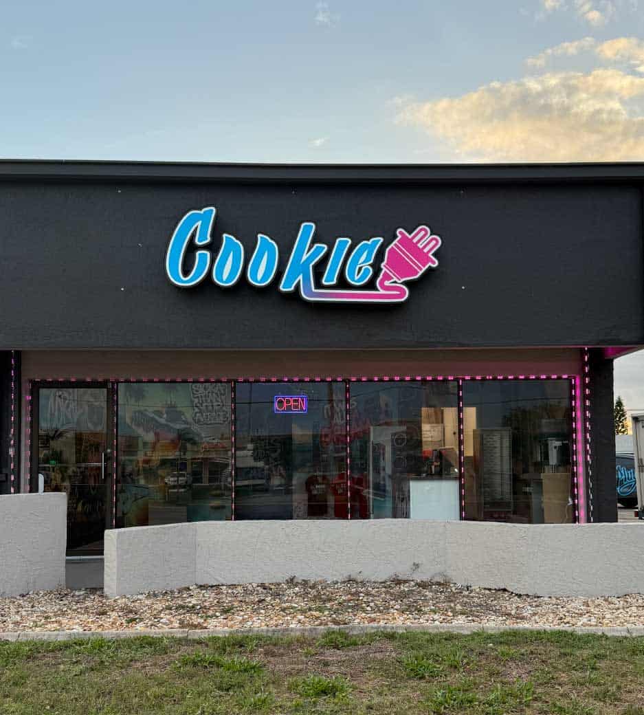 black storefront with a pink, purple, and blue gradient sign that says 'Cookie' with an electrical plug attached to it with graffiti and an alligator in the window.