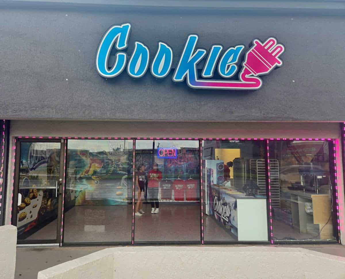 Gray storefront with a pink, purple, and blue gradient sign that says 'Cookie' with an electrical plug attached to it with graffiti and an alligator in the window.