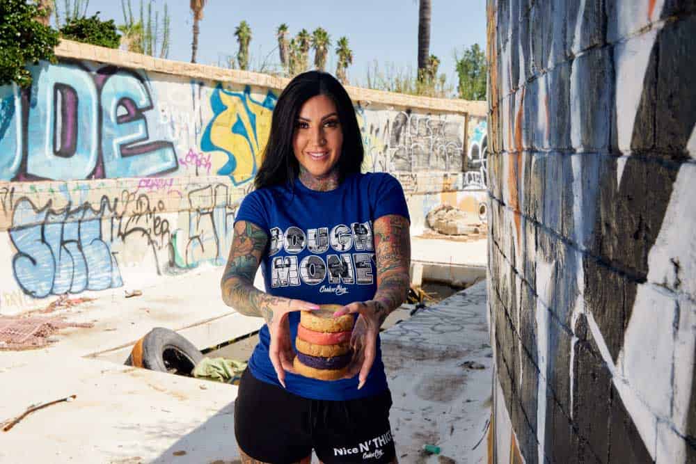 Girl with black hair in a blue shirt that is hold a stack of cookies in various flavors like purple velvet, strawberry cheesecake, and cereal flavored who is standing in an alleyway covered in graffiti
