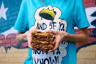 Girl with blue graffiti shirt on with a crown who is hold thick stacks of chocolate chip cookies.