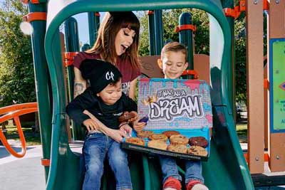 Women sitting on a kid's slide with two boys who are holding a large box of cookies that says 'it was all a dream' in graffiti writing.