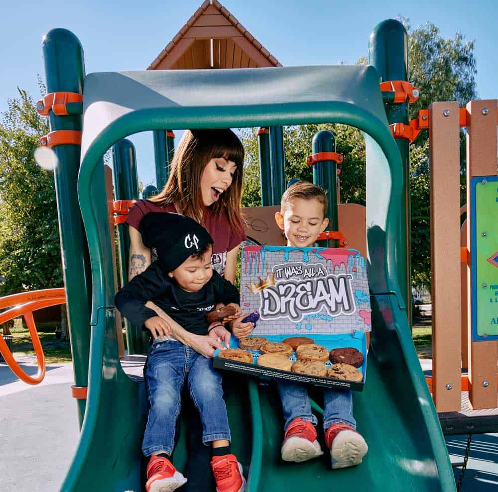 Women sitting on a kid's slide with two boys who are holding a large box of cookies that says 'it was all a dream' in graffiti writing.