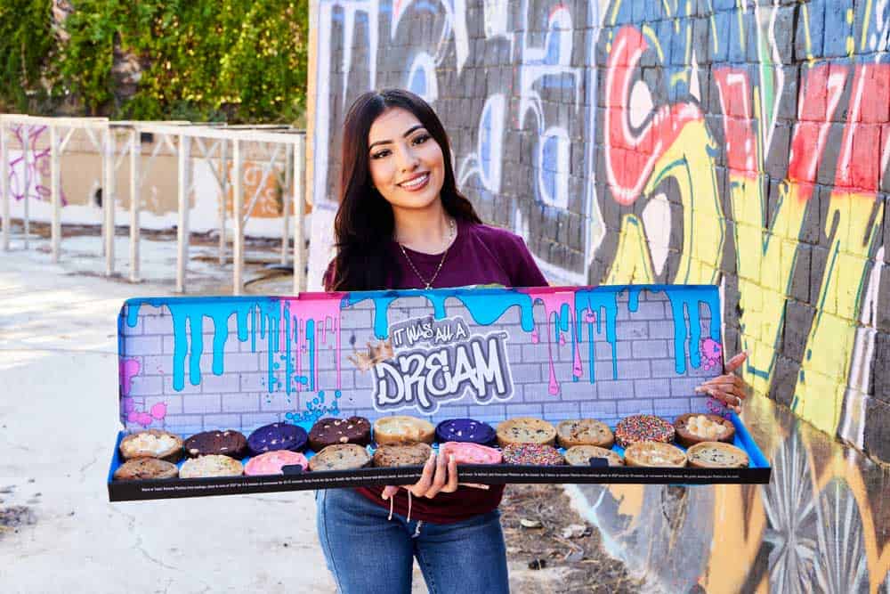Smiling girl holding box that's covered in color graffiti with large cookies in various flavors