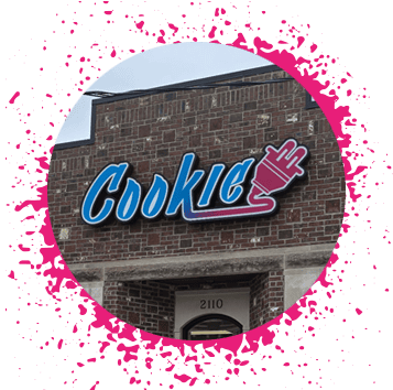 Blue, pink, and purple gradient sign says 'Cookie Plug' over a brick storefront with large windows with a hot pink spray paint border