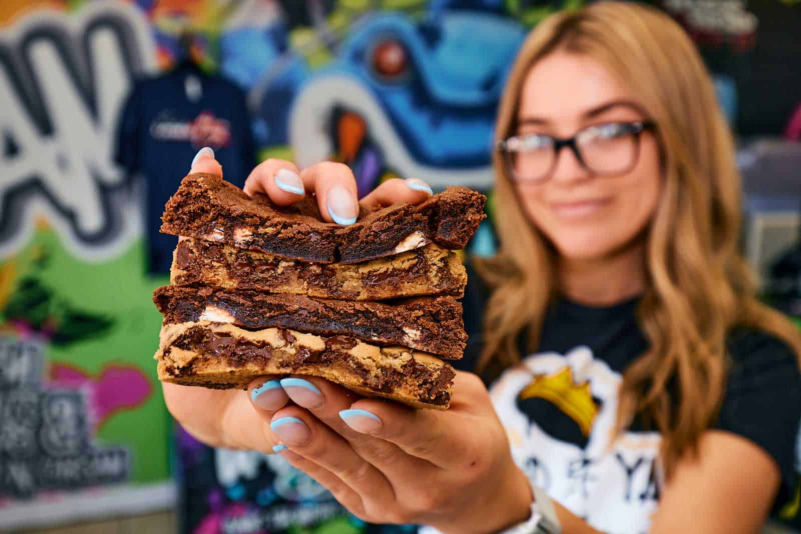 blurred out girl in front of a graffiti colored wall who has a thick stack of 4 cookies that are sliced down the center with melted while and dark chocolate chips