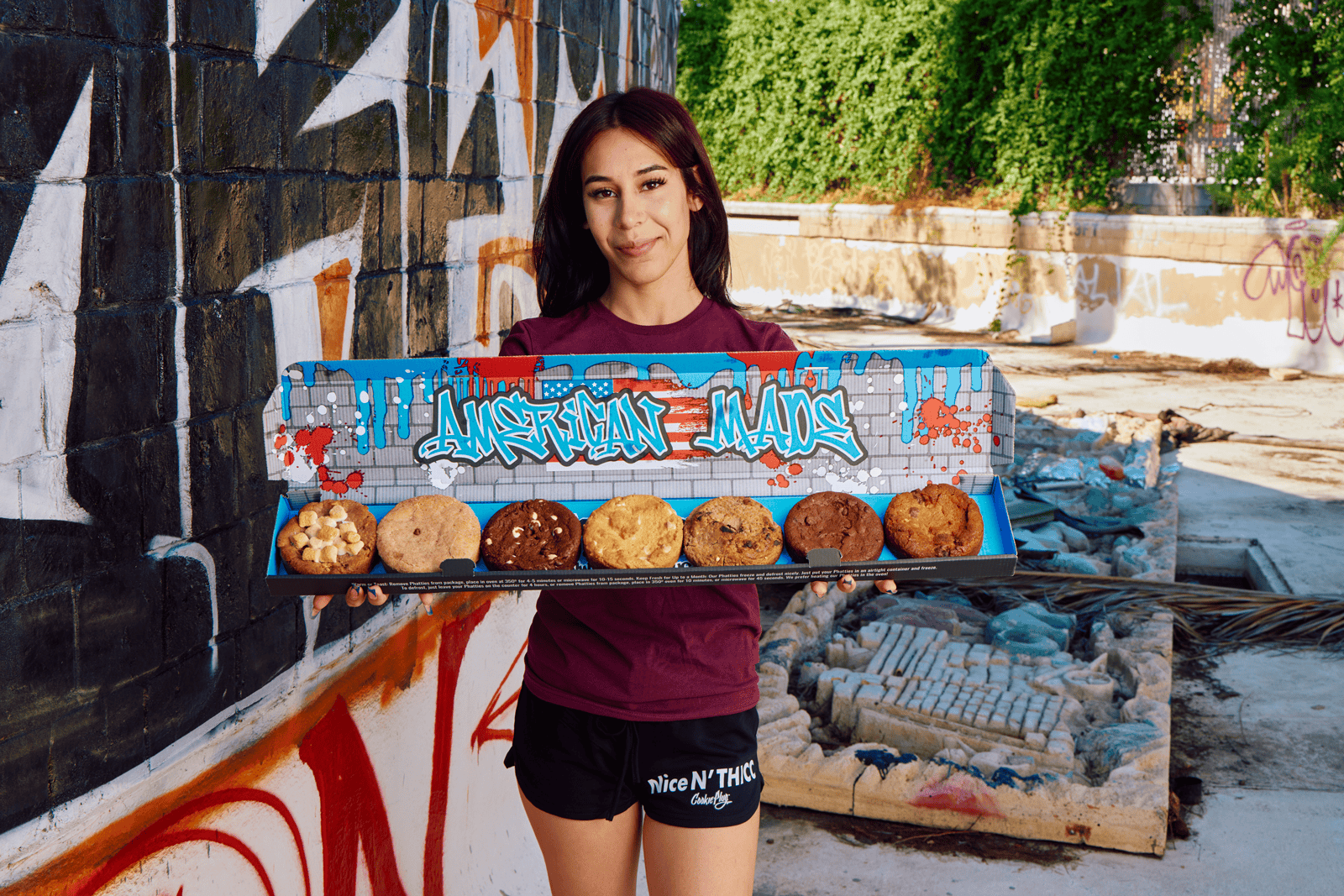 Girl holding a large cookie box that says 'American Made' in blue, white, and black graffiti letters with blue and red paint splotches. The box holds 7 cookies in various flavors, such as chocolate with white chocolate chips, chocolate chip cookie, cinnamon cookies, and smores cookies
