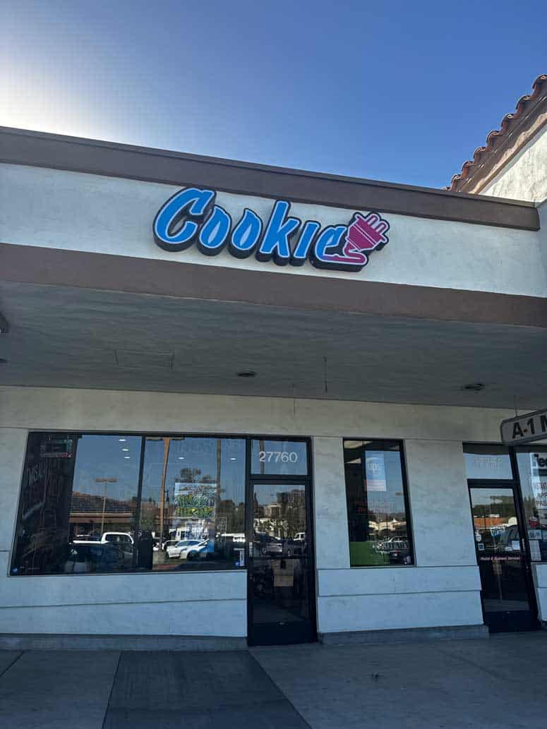 Beige storefront with a large blue, purple, and pink gradient sign that says 'Cookie' with an electrical plug attached to the end in graffiti letters over a window sign that says 'Now Hiring'.