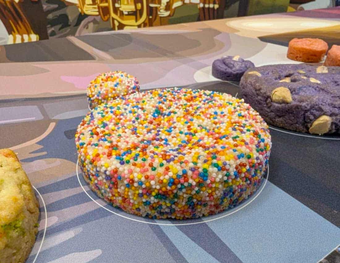 Large sugar cookie with sprinkles all over the top sits upon a counter of a cookie shop display.