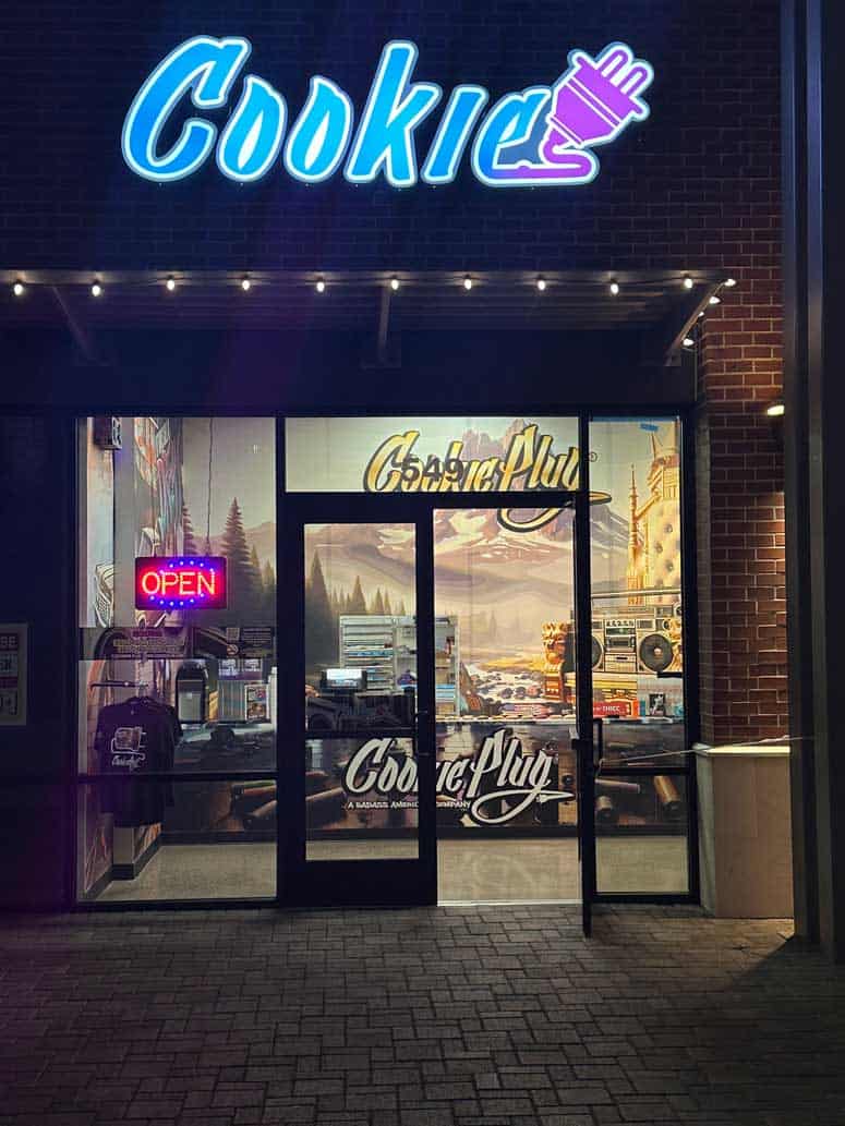 Brick store front with a golden yellow sign that says 'Cookie Plug' with cookie racks in the window and a graffiti boombox.