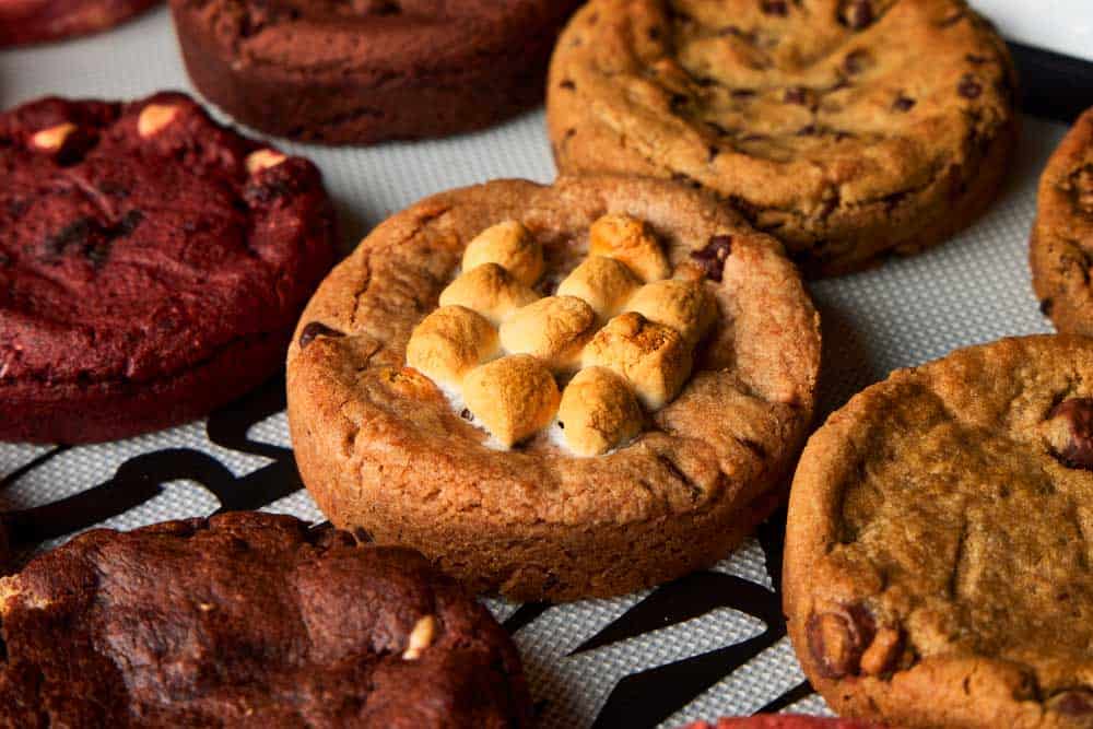 a picture of large, thick cookies on a baking tray that contains a roasted marshmallow and chocolate, read velvet cake cookie, and peanut butter cookie.