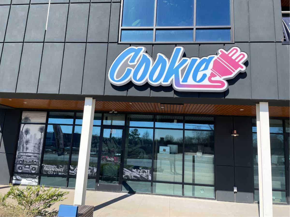 Storefront with large glass windows with a white, blue, purple, and pink sign that says 'Cookie' with an electrical plug attached to it.
