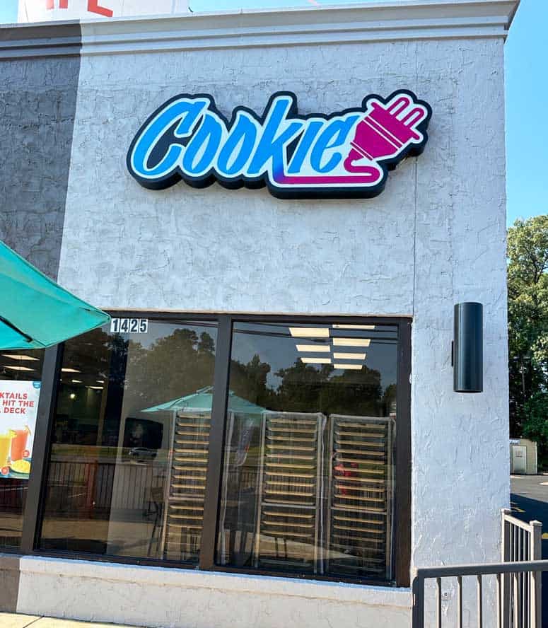 gray stone storefront with a blue, purple, and pink sign in graffiti type that says 'Cookie' with an electrical plug attached to it with cookie baking racks in the window.